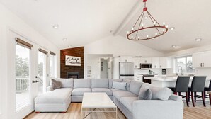 Uma sala de estar moderna com um sofá seccional, uma área de jantar com mesa e cadeiras, e uma cozinha ao fundo.