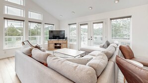 Uma sala de estar ampla com um sofá seccional grande, uma mesa de centro e uma TV de tela plana em um suporte de madeira. Janelas amplas oferecem luz natural e vista para o exterior.