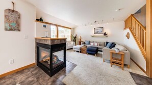 Ein modernes Wohnzimmer mit Kamin, einer Sofagarnitur, einer Holztreppe und einem großen Fenster.