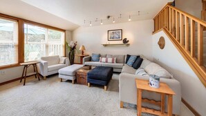 Ein gemütliches Wohnzimmer mit einer Couch, Hocker und einem kleinen Holztisch. Rechts befindet sich eine Treppe und links ein Fenster mit Jalousien.
