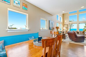 Ein geräumiges Wohnzimmer mit einem großen Esstisch aus Holz, blauen Polstersitzgelegenheiten und zahlreichen Fenstern mit Blick auf den Ozean.