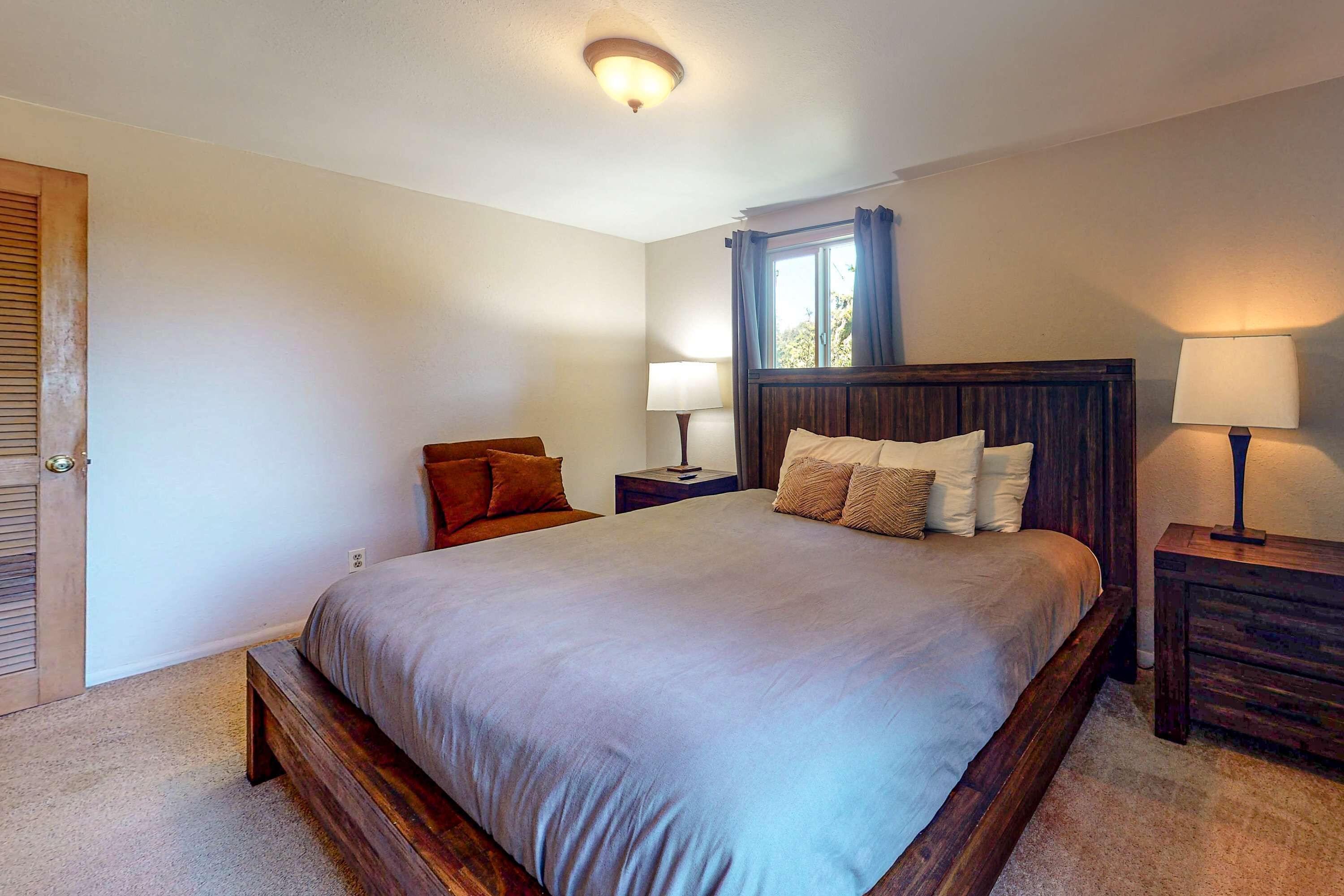 A bedroom with a large bed, two bedside tables with lamps, a window with curtains, and a door with a wooden frame.