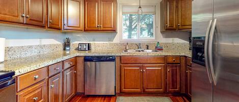 A kitchen with wooden cabinets, stainless steel appliances, granite countertops, and a window with a view of trees.