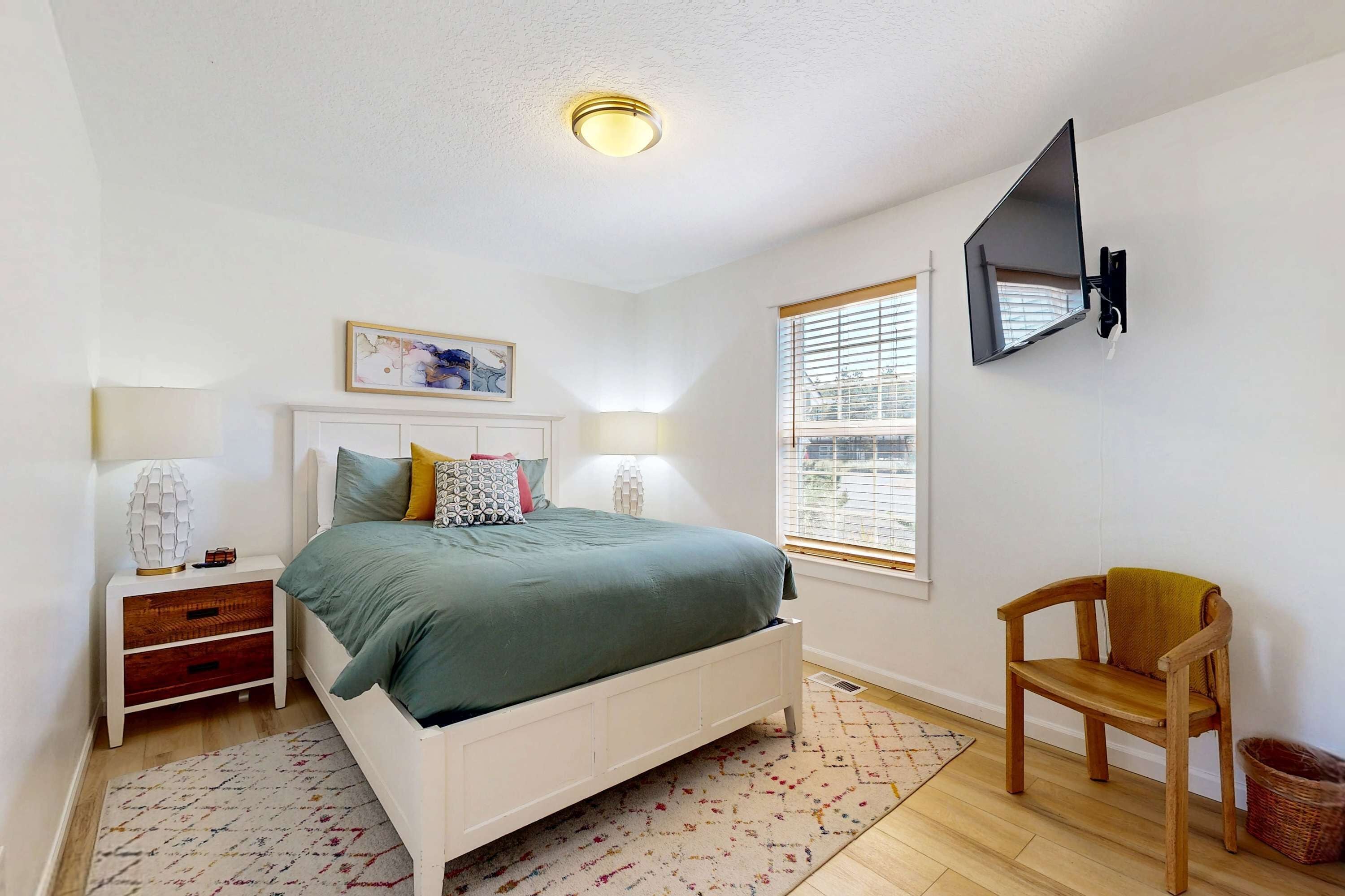 A bedroom with a bed, bedside table, lamp, chair, and a mounted TV.