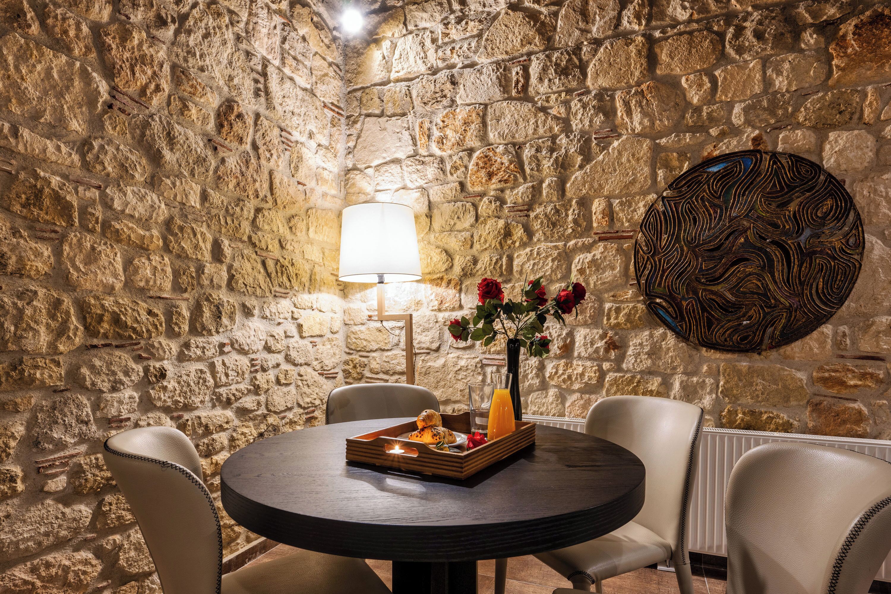 A stone-walled room with a round table, chairs, a lamp, and a decorative plate.