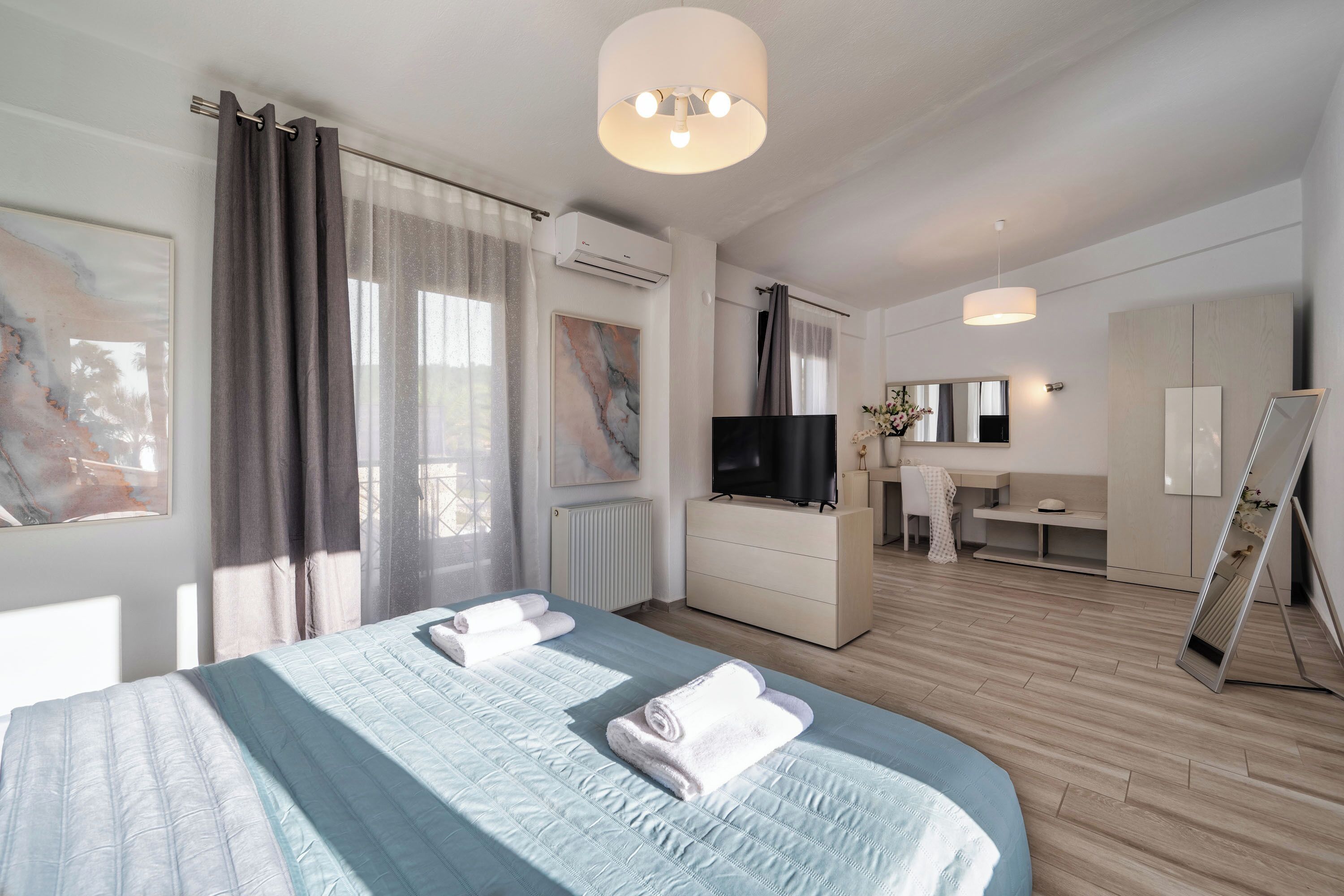 A modern hotel room with a large bed, a flat-screen TV, a desk with a chair, and a large mirror.