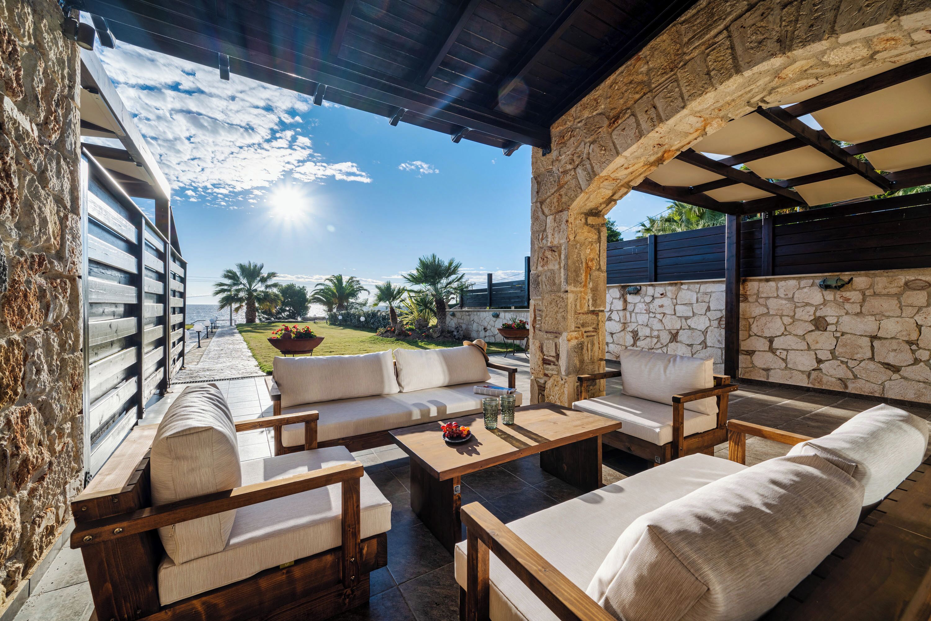 Eine Terrasse im Freien mit Holzmöbeln, Steinmauern und Blick auf Palmen und einen Garten.