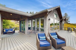 A covered outdoor deck with wicker furniture and glass doors leading to a house. - Luxury Coastal Retreat | Wrap Around Deck w/Panoramic Ocean Views (Pacific City)