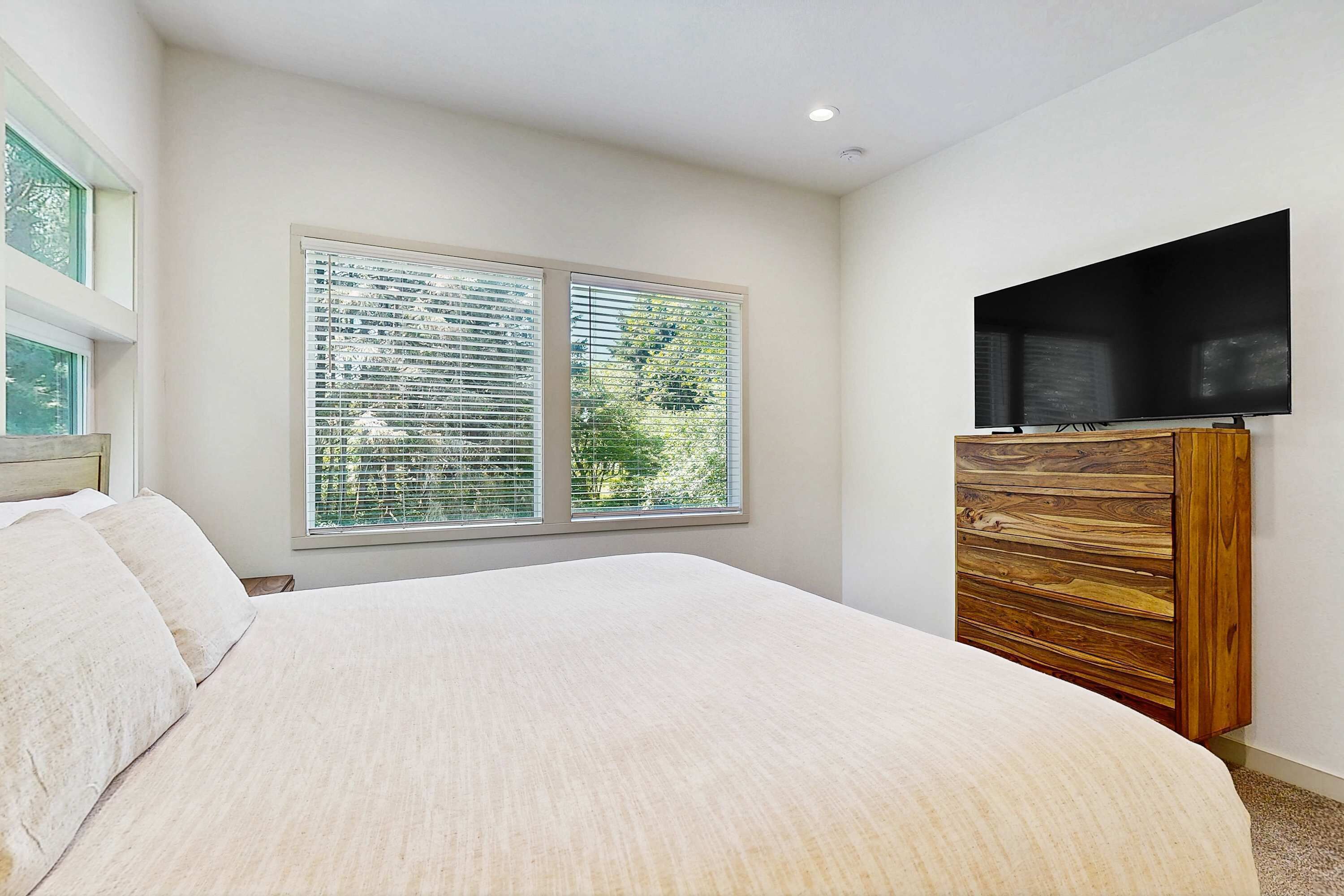 A bedroom with a large bed, a TV mounted on a wooden cabinet, and a window with blinds.