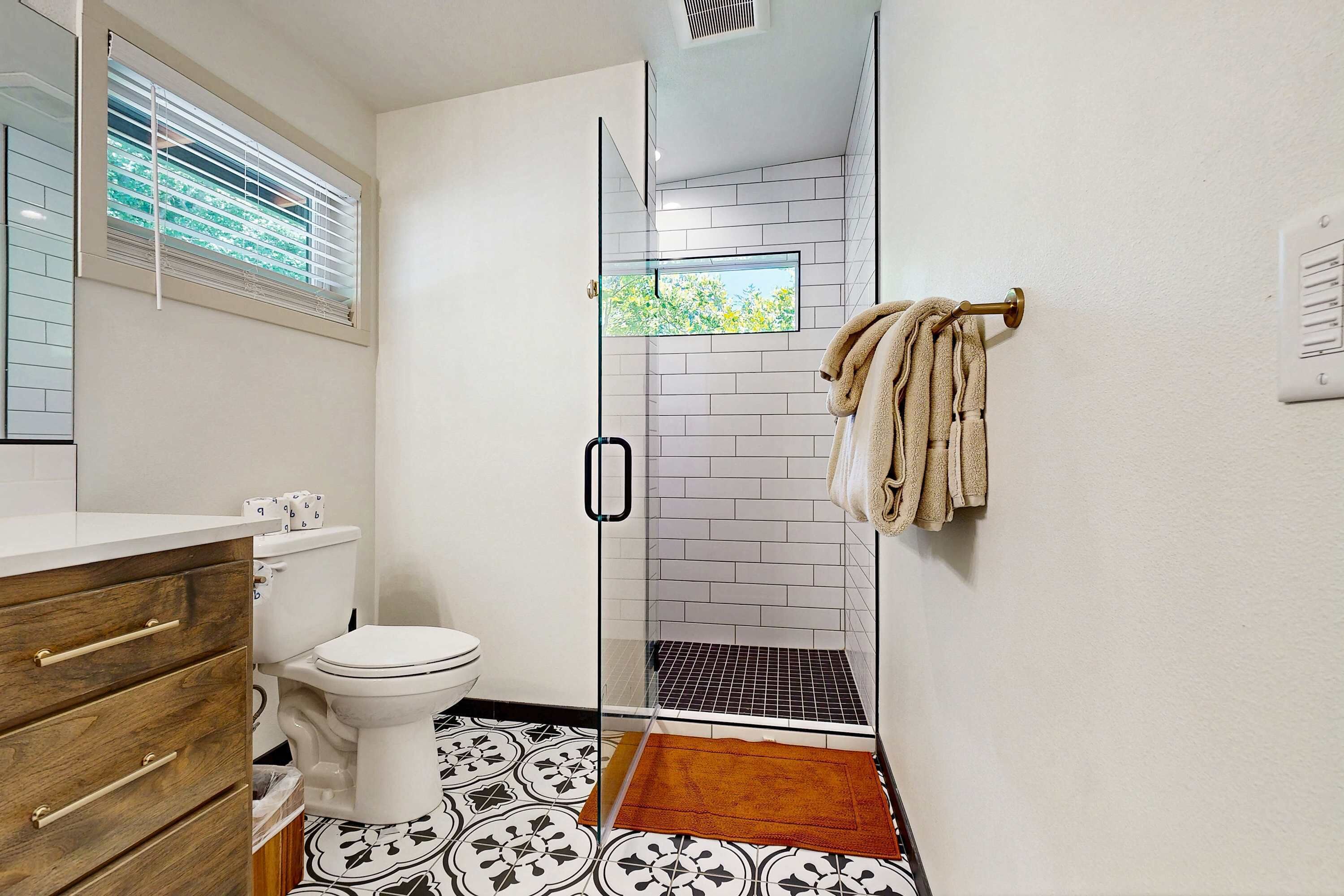 Un baño con inodoro, un mueble de madera, cabina de ducha con mampara de cristal y suelo estampado.
