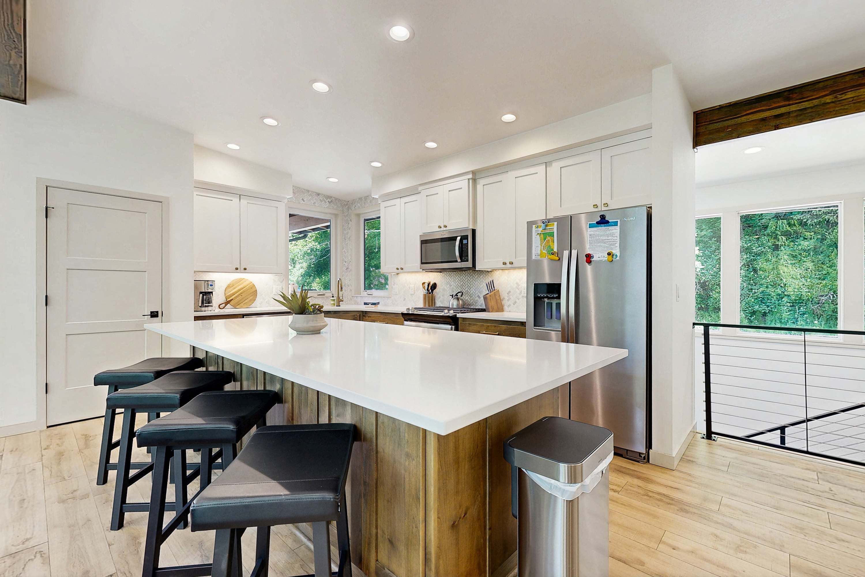A modern kitchen with white cabinets, a large island with bar stools, stainless steel appliances, and a view of greenery through large windows.