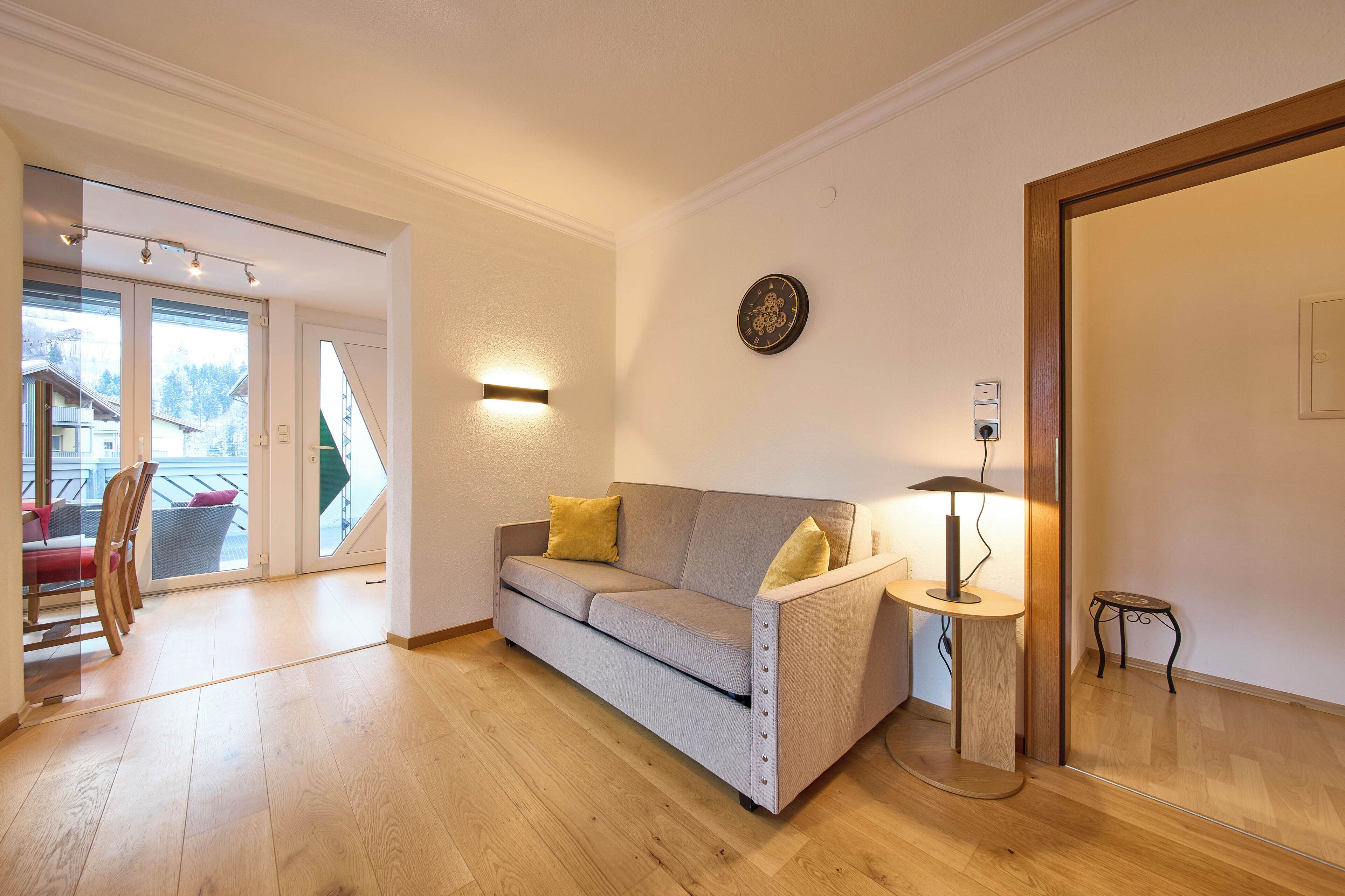 A modern living room with a grey sofa, wooden flooring, and a small side table with a lamp.