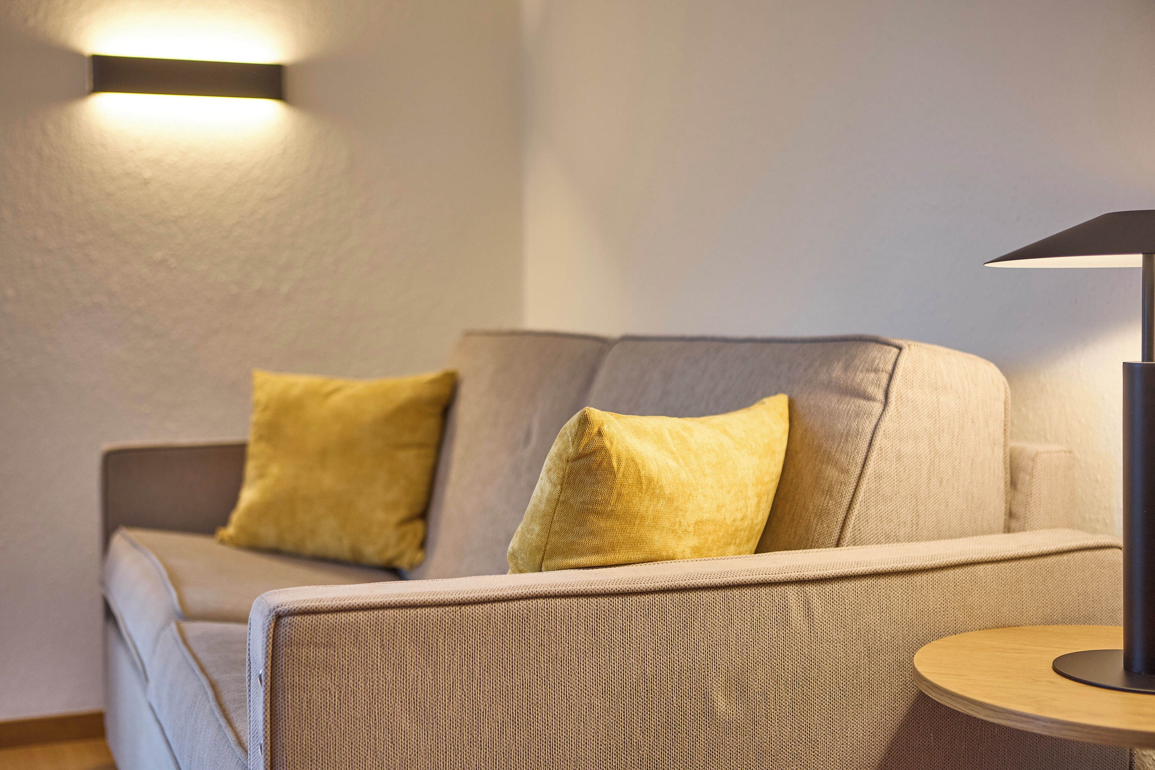 A modern living room with a sofa and two yellow cushions, a round wooden side table, and a floor lamp.