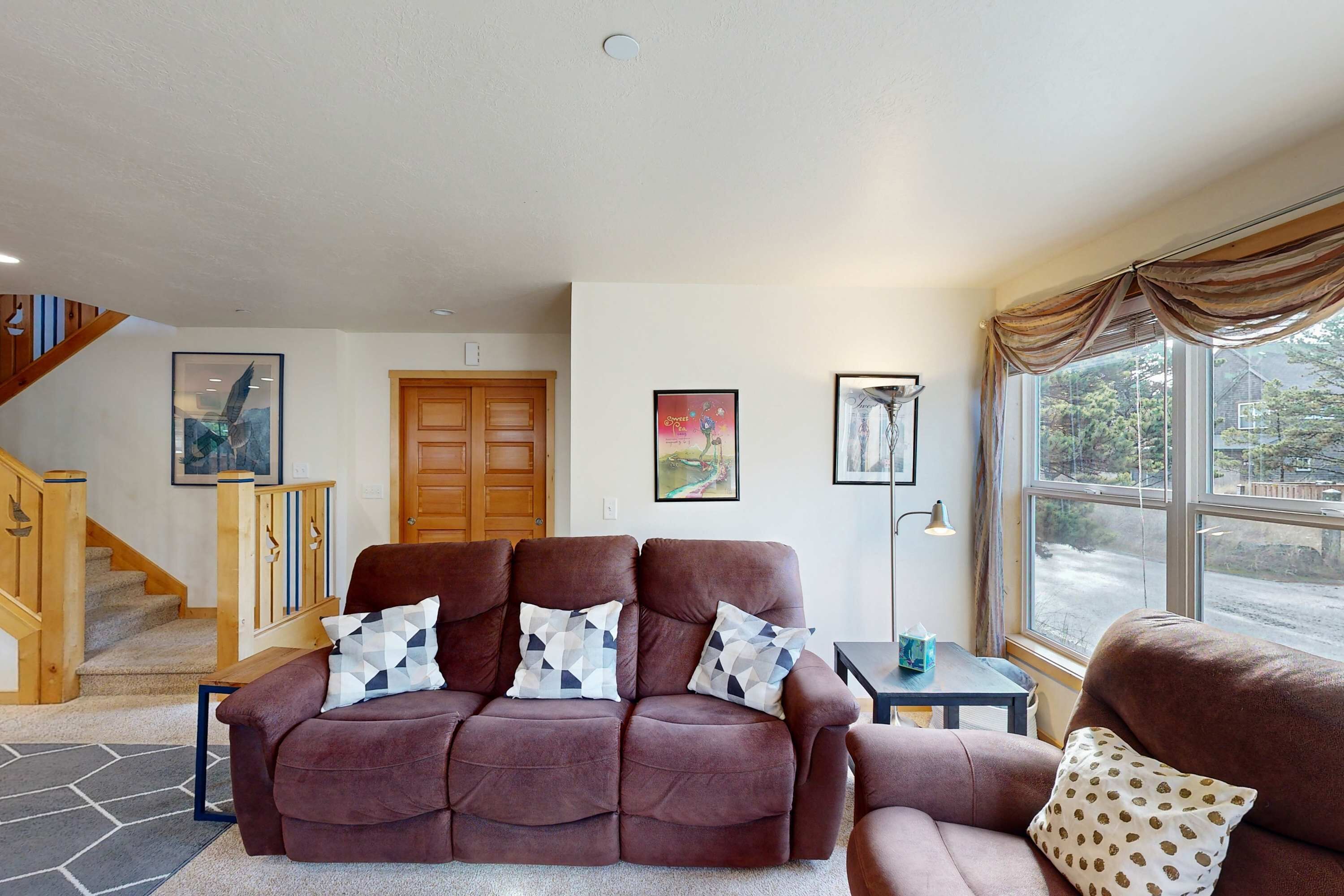 A living room with a brown sofa set, a wooden staircase, and a large window with curtains.
