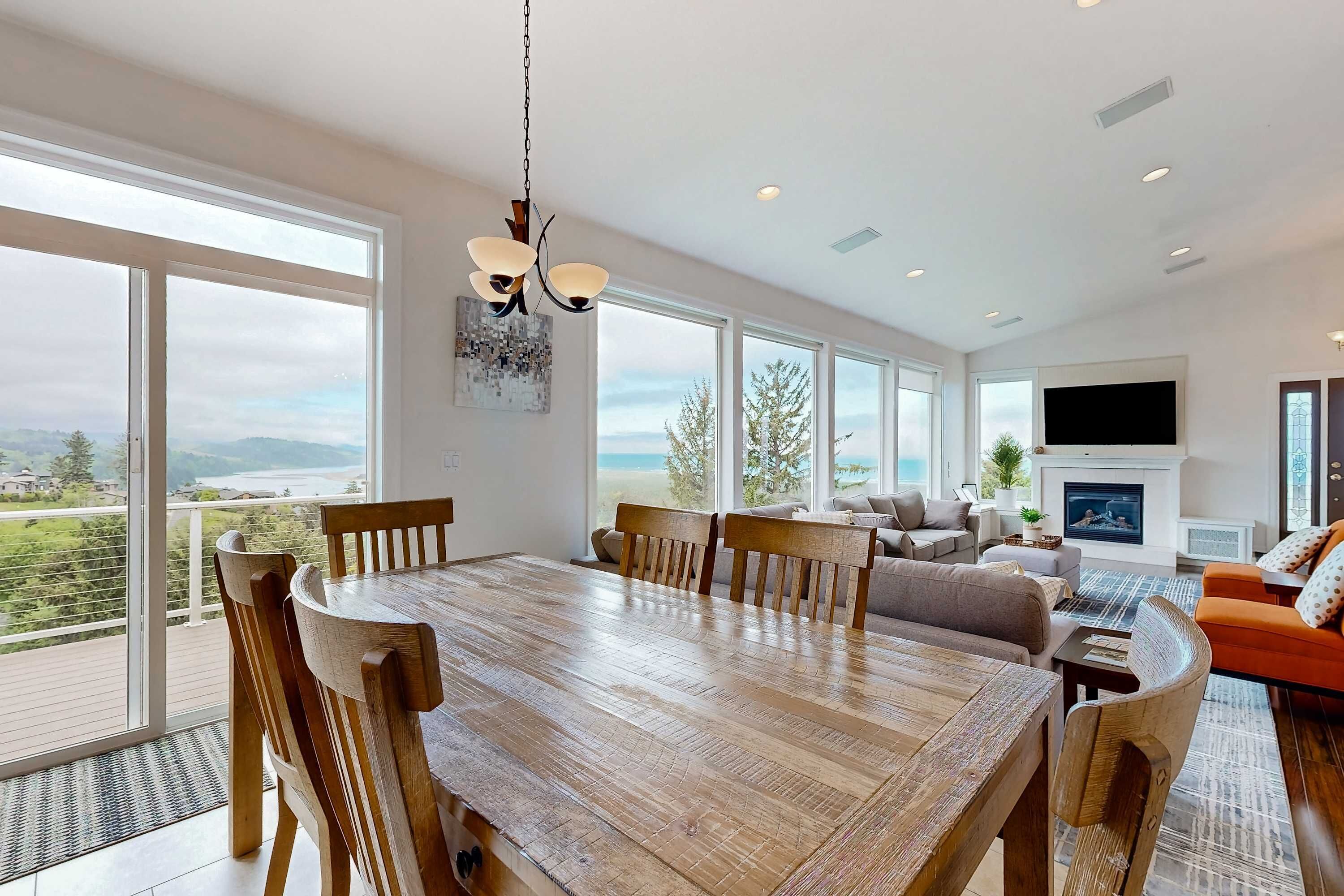 A spacious living room with a large wooden dining table, comfortable seating, a fireplace, and large windows offering a scenic view.