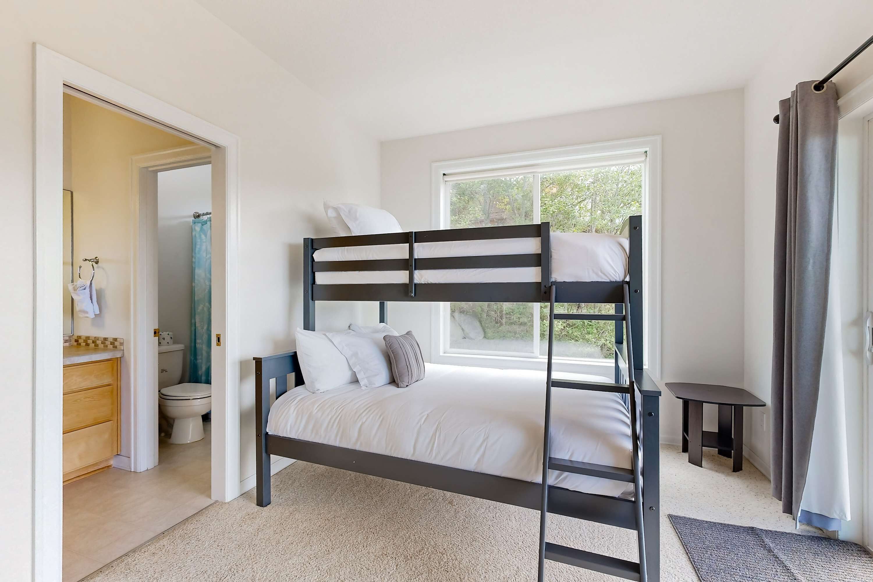 Un lit superposé dans une pièce avec une fenêtre, une petite table et une salle de bain visible à travers une porte ouverte.