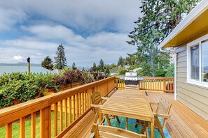 Une terrasse en bois avec une table et des chaises, offrant une vue sur un plan d’eau et des montagnes.