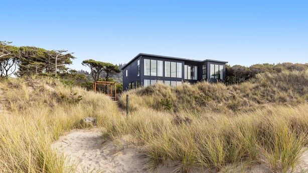 Une maison moderne aux grandes fenêtres, entourée de dunes de sable et d’une végétation clairsemée.
