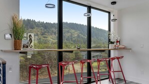 Une pièce moderne avec de grandes fenêtres, un comptoir de bar en bois, des tabourets de bar rouges et une vue sur une colline boisée.