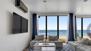 Un salon moderne doté d’un grand téléviseur, d’une table basse blanche et d’un canapé placé face à une vue sur la plage, visible à travers de grandes fenêtres.