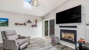 A modern living room with a gray sofa, a flat-screen TV, a fireplace, and a large window. - Home with gas fireplace washer dryer and 2 outdoor spaces walk to beach (Tierra Del Mar)