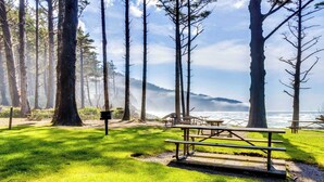 Ein Picknickplatz mit Bänken und einem Picknicktisch, umgeben von hohen Bäumen und Blick auf das Meer.