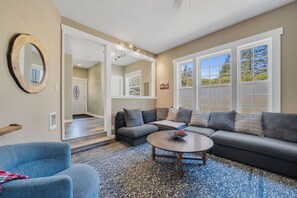 A living room with a sofa, a coffee table, and a blue armchair. There is a large window with multiple panes and a view of trees. - Cozy Coast Escape, Close to the Beach, Family Friendly (Netarts)