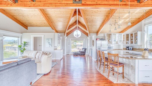 A spacious kitchen with wooden beams, a dining area with bar stools, and a living room with a sofa and a large window. - Elegant home with sauna private hot tub and ocean views accessible by elevator (Cape Meares)