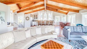A spacious living area with a high wooden ceiling, a kitchen in the background, and a dining area with chairs. - Elegant home with sauna private hot tub and ocean views accessible by elevator (Cape Meares)