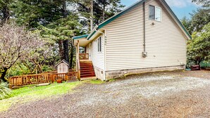 A small, single-story house with a porch and a small shed, surrounded by trees and a gravel driveway. - Renovated three story home by the beach with a furnished deck and washer dryer (Cape Meares)
