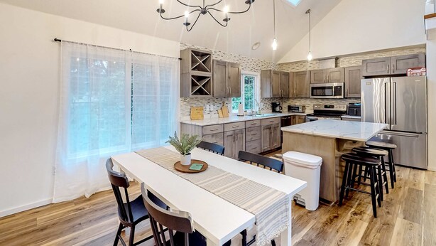 A modern kitchen with a central island, wooden cabinets, stainless steel appliances, and a dining area with a white table and chairs. - Renovated three story home by the beach with a furnished deck and washer dryer (Cape Meares)