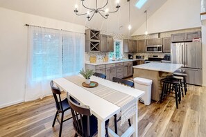 A modern kitchen with a central island, wooden cabinets, stainless steel appliances, and a dining area with a white table and chairs. - Renovated three story home by the beach with a furnished deck and washer dryer (Cape Meares)