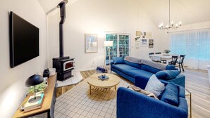 A modern living room with a blue sofa, a round coffee table, a fireplace, and a dining area with a table and chairs. - Renovated three story home by the beach with a furnished deck and washer dryer (Cape Meares)