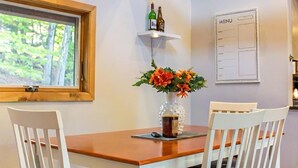 Un comedor con una mesa de madera, sillas blancas con asientos acolchados, un jarrón con flores, un menú en la pared y una ventana con vistas a árboles.