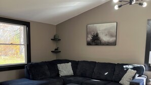 Una sala de estar moderna con un sofá seccional, un reposapiés a juego, un ventanal con vistas a árboles, una lámpara de techo e imagen enmarcada en la pared.