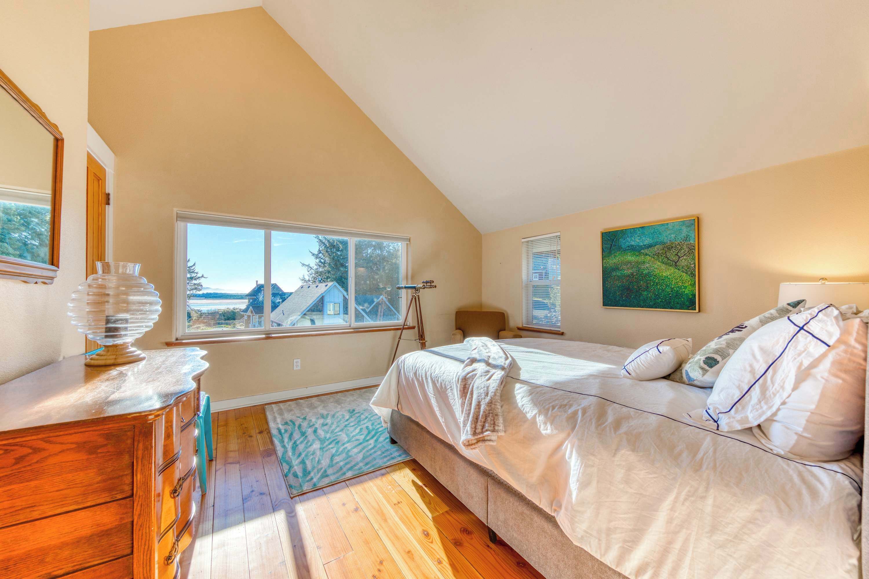 A bedroom with a large bed, a wooden dresser, a window with a view, and a painting on the wall.