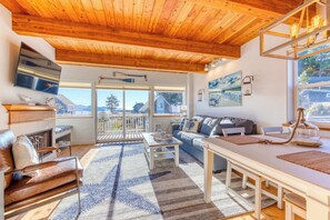 Amplia sala de estar con techo de madera, chimenea, sofá, mesa de comedor y un ventanal con vistas al exterior.