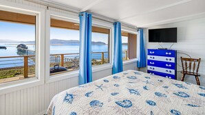 A bedroom with a large bed, a dresser, a chair, and a TV. There are large windows with curtains, and a view of the water and mountains. - Gorgeous home overlooking the bay near a boat launch dog friendly (Garibaldi)