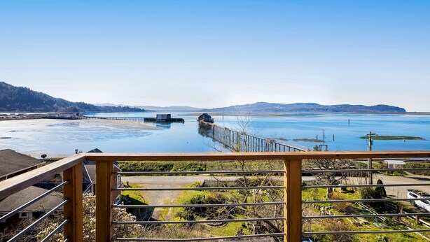 A coastal view from a balcony with a wooden railing, a dock extending into the water, and a distant shoreline with hills. - Gorgeous home overlooking the bay near a boat launch dog friendly (Garibaldi)