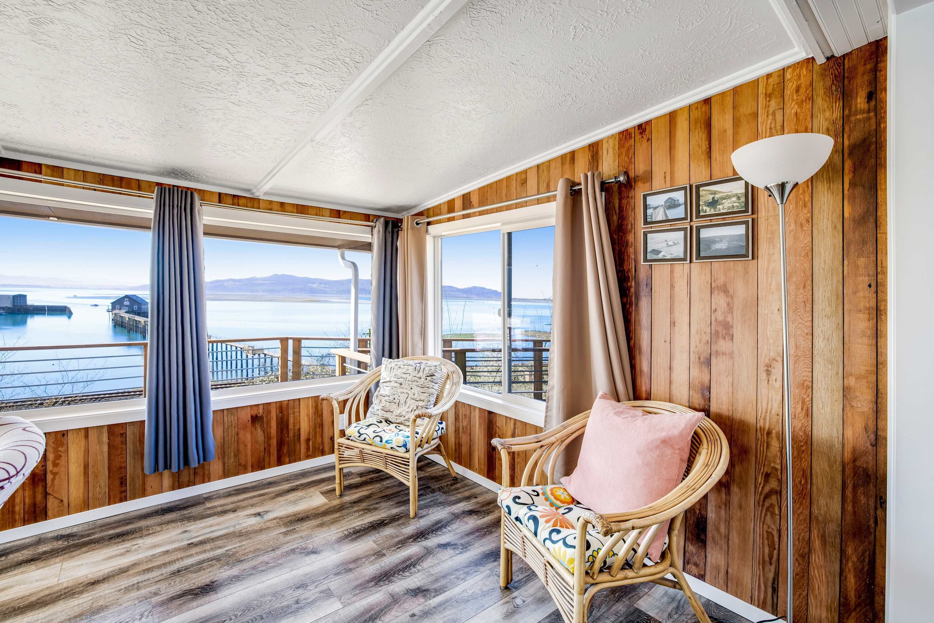 A room with wooden walls and a view of a body of water. There are two wicker chairs with cushions, a floor lamp, and a window with curtains.