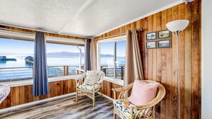A room with wooden walls and a view of a body of water. There are two wicker chairs with cushions, a floor lamp, and a window with curtains. - Gorgeous home overlooking the bay near a boat launch dog friendly (Garibaldi)