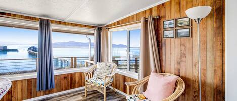A room with wooden walls and a view of a body of water. There are two wicker chairs with cushions, a floor lamp, and a window with curtains.