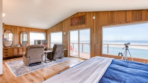 Ett sovrum med en stor säng, ett skrivbord, två stolar, en spegel och utsikt över havet genom stora fönster.