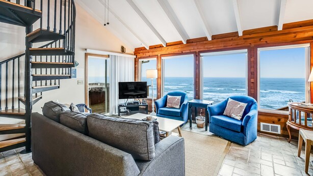 Amplia sala de estar con un sofá grande, un sillón azul y un televisor. Se ve una escalera de madera y vista al océano.