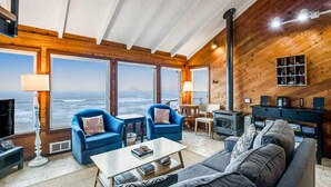 Una sala de estar acogedora con un gran ventanal con vista al océano, paredes de madera, una chimenea y asientos cómodos.