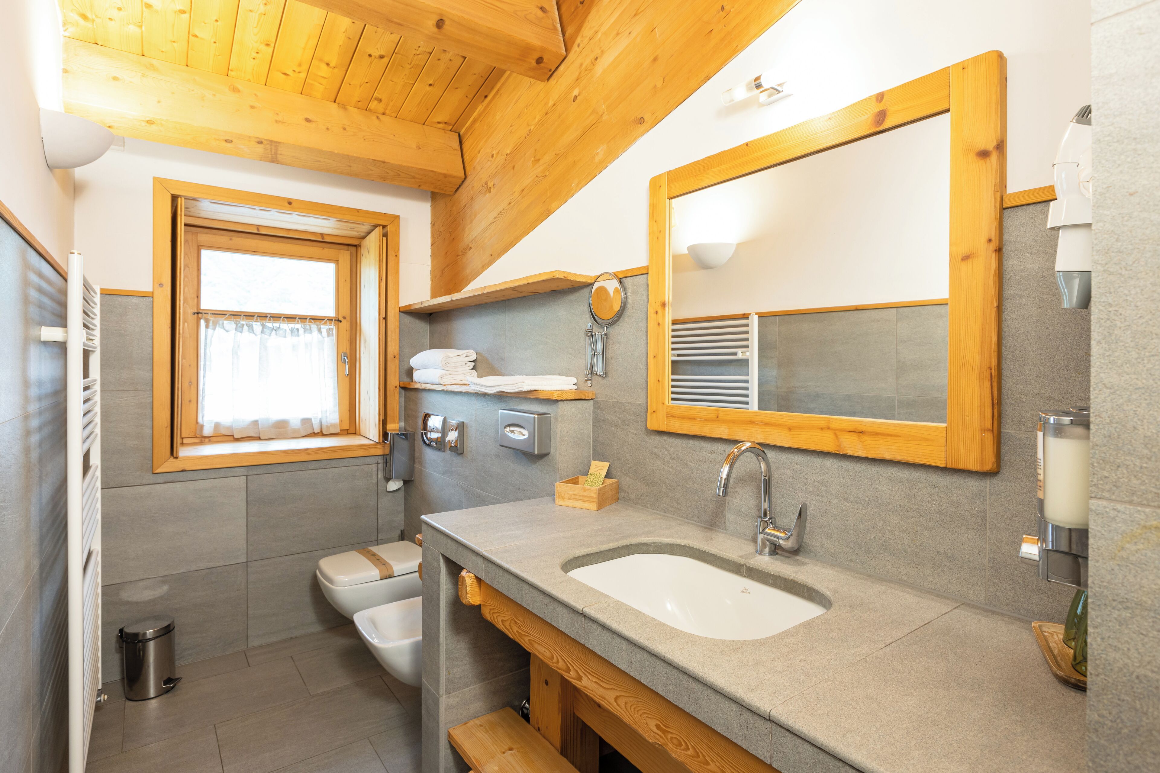 A bathroom with a wooden ceiling, a window with curtains, a sink, a mirror, and a toilet.
