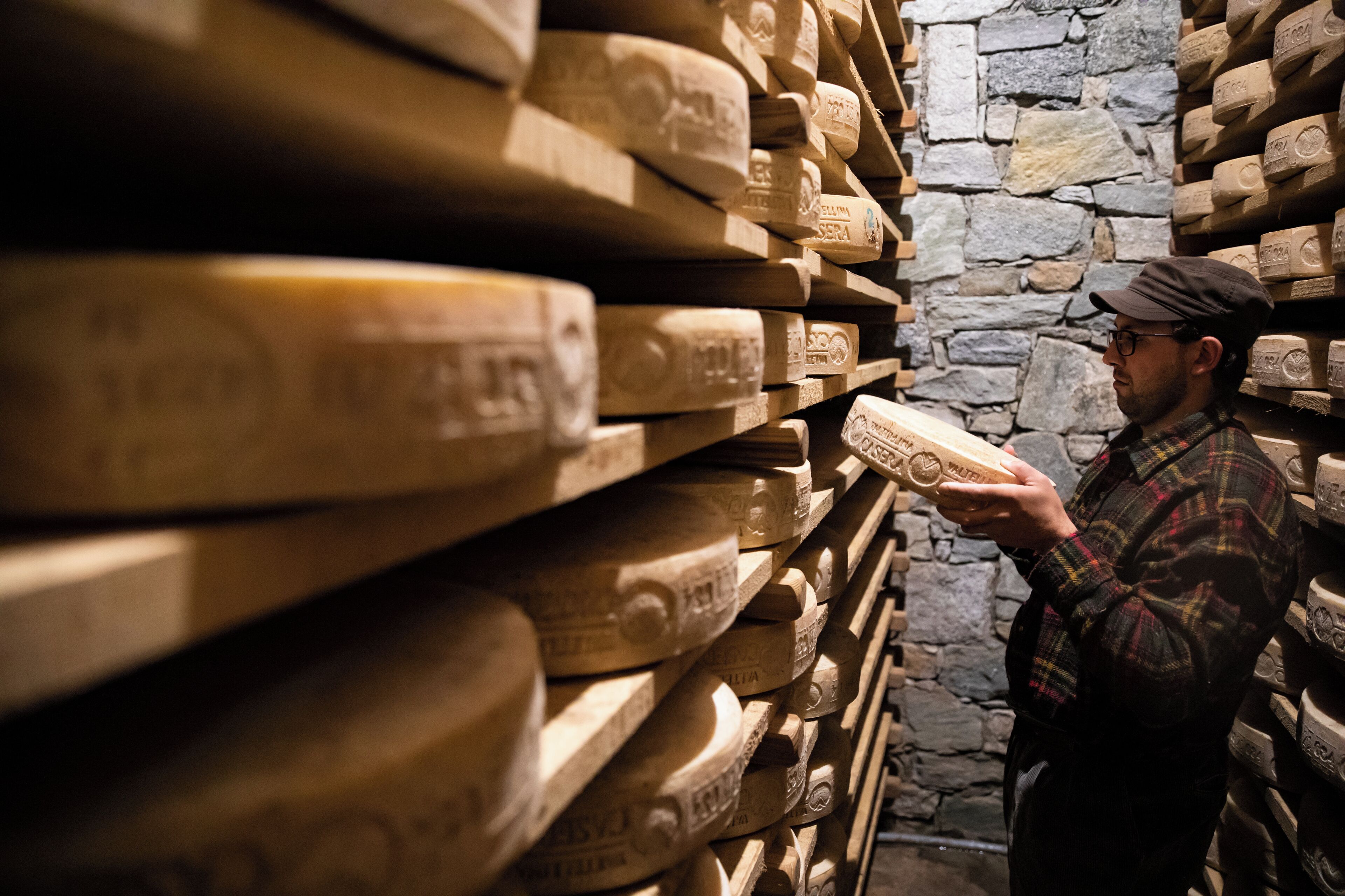 A person examining cheese wheels in a cellar with stone walls.