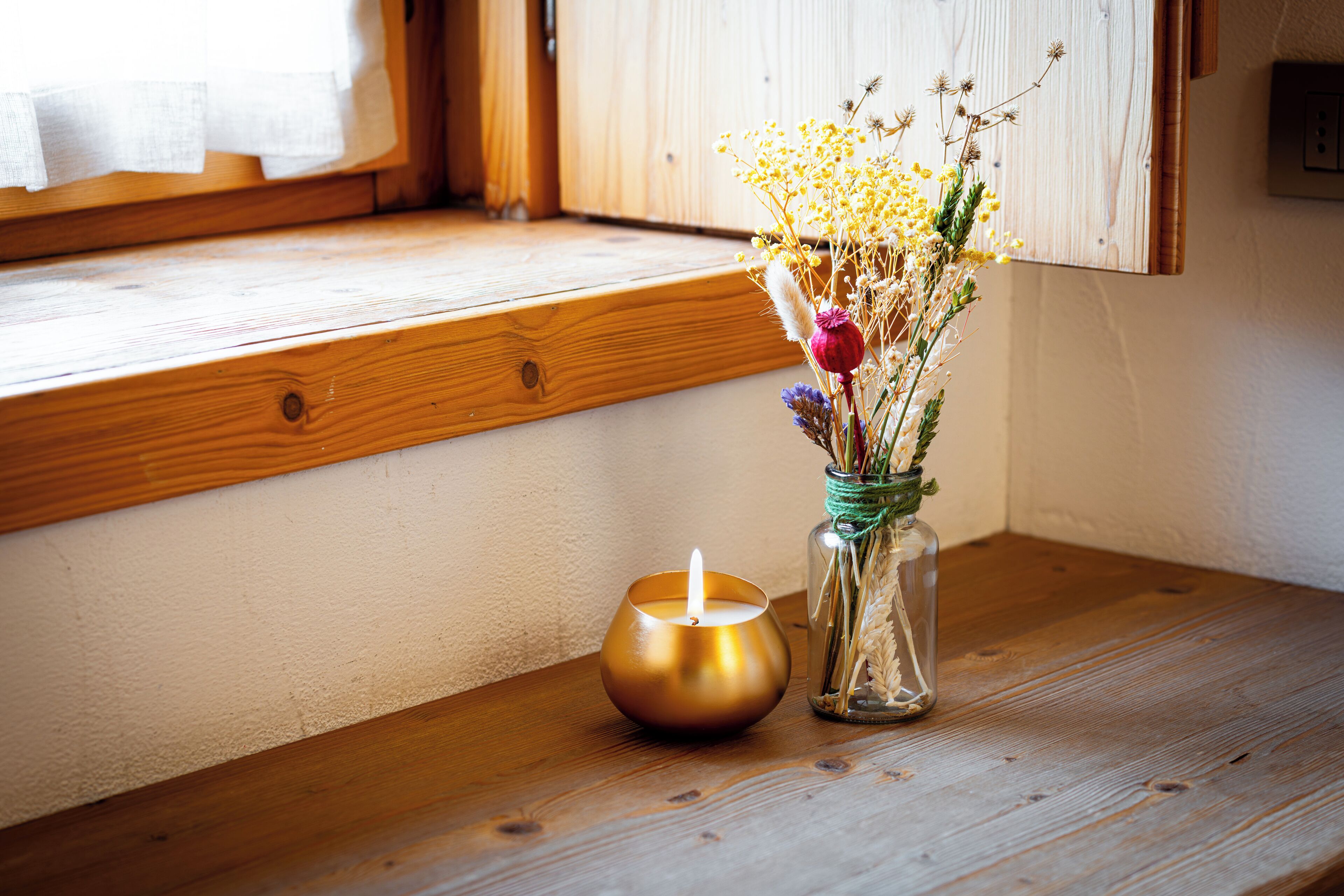 A candle and a vase with dried flowers on a wooden table near a window.