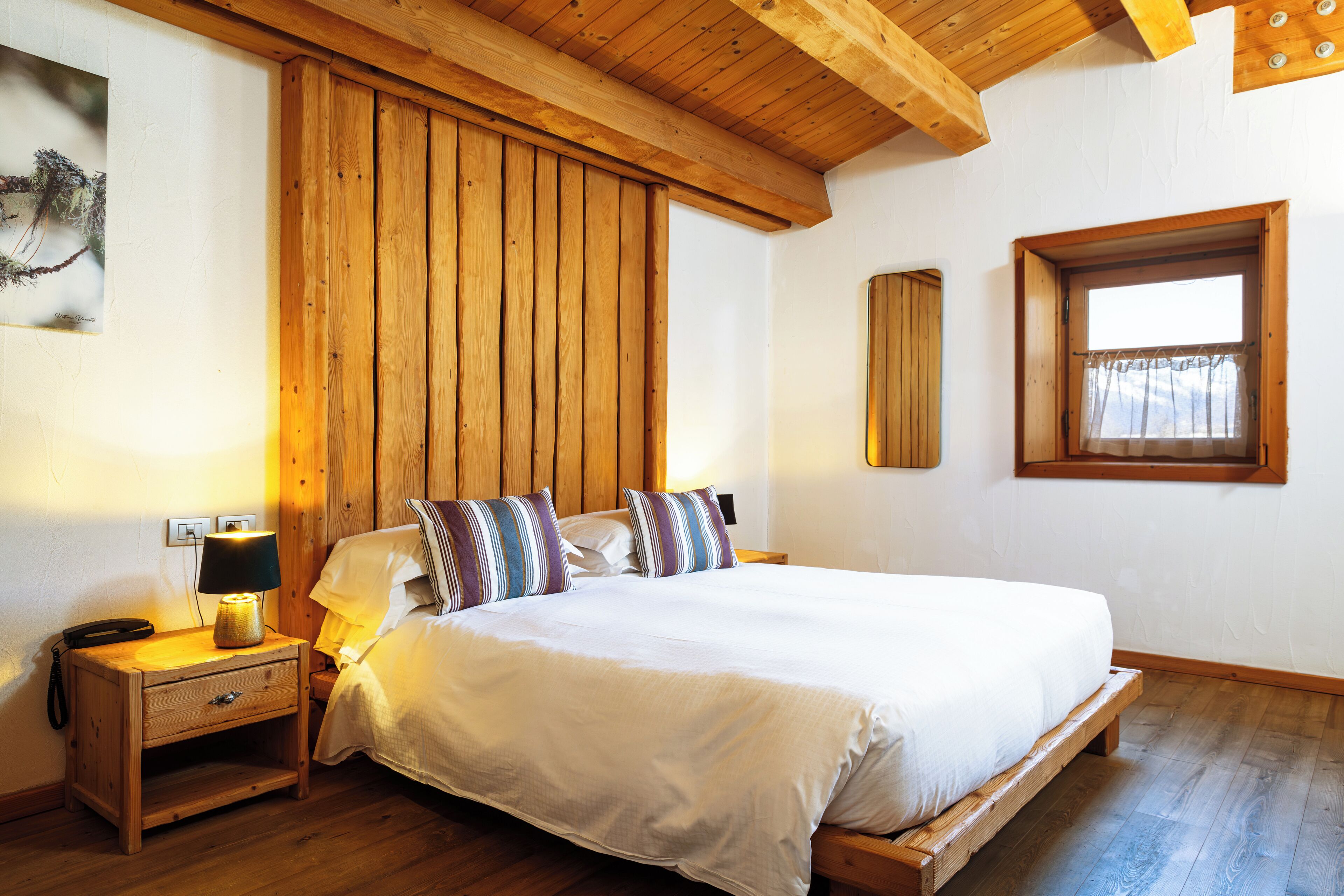A bedroom with a wooden bed frame, white bedding, a bedside table with a lamp, and a window with curtains.