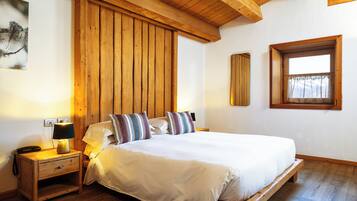 Un dormitorio con estructura de cama de madera, ropa de cama blanca, una mesita de noche con lámpara y una ventana con cortinas.
